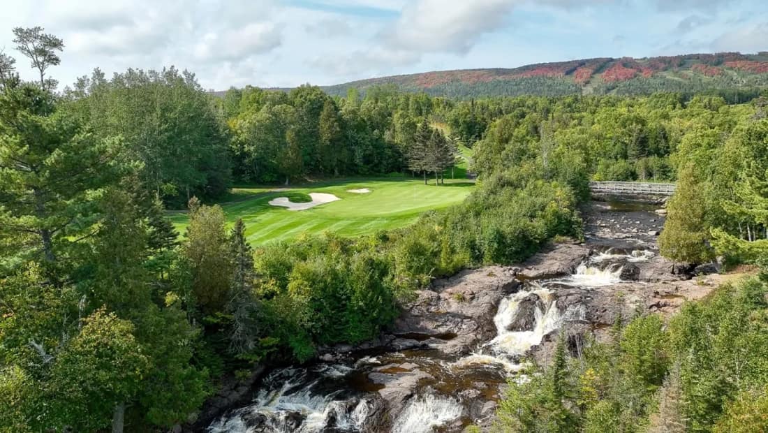 A golf green framed by north-shore forest with a waterfall in the foreground